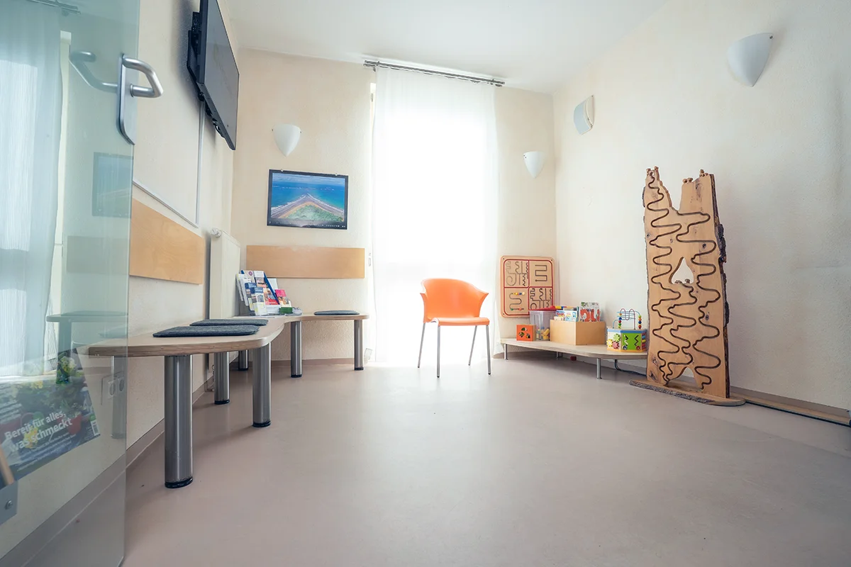 Helles Wartezimmer der Arztpraxis mit Sitzbank, Stuhl und Spielecke für Kinder.